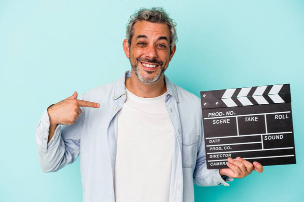 Middle age caucasian man holding a clapperboard isolated on blue background  person pointing by hand to a shirt copy space, proud and confident - Foto, Imagen