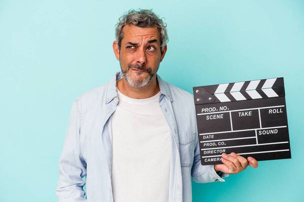 Middle age caucasian man holding a clapperboard isolated on blue background  confused, feels doubtful and unsure. - Photo, Image