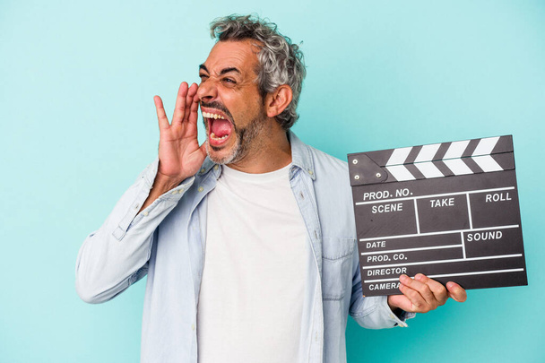 Middle age caucasian man holding a clapperboard isolated on blue background  shouting and holding palm near opened mouth. - Foto, Imagen
