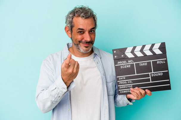 Middle age caucasian man holding a clapperboard isolated on blue background  pointing with finger at you as if inviting come closer. - Foto, Imagen