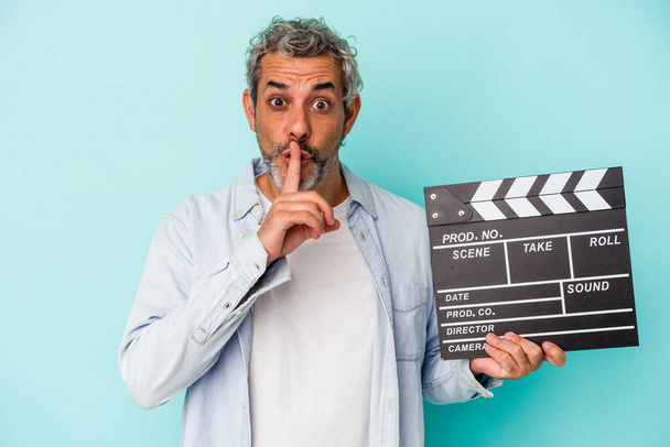 Middle age caucasian man holding a clapperboard isolated on blue background  keeping a secret or asking for silence. - Photo, Image