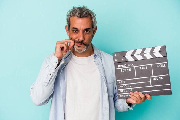 Middle age caucasian man holding a clapperboard isolated on blue background  with fingers on lips keeping a secret. - Foto, Imagen