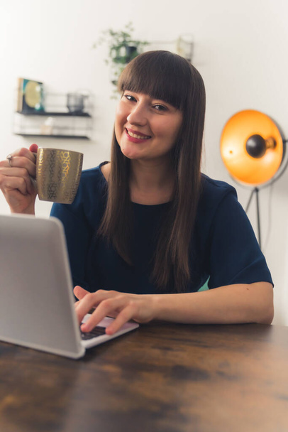Vertikální záběr evropské dlouhosrsté brunetky, jak se dívá do kamery, drží hrnek s čajem a pomocí notebooku. - Fotografie, Obrázek