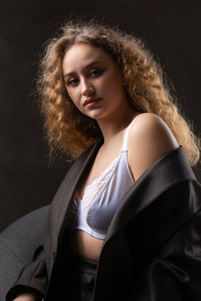 Portrait of a young red-haired curly sexy girl in a jacket with long hair in the studio. Shows off her curvaceous breasts in a bra. - Foto, Bild