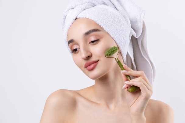 Beautiful girl with thick eyebrows and perfect skin at white background, towel on head, beauty photo. Holds a cosmetic roller. - Foto, Bild