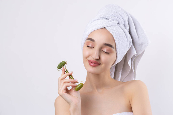 Beautiful girl with thick eyebrows and perfect skin at white background, towel on head, beauty photo. Holds a cosmetic roller. - Zdjęcie, obraz
