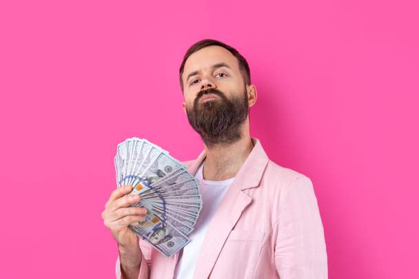 Portrait of a contented young businessman with a beard dressed in a pink jacket showing us dollar banknotes against a red studio background. Taste, smell of money. - Fotografie, Obrázek