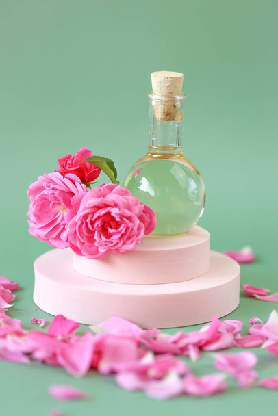 Olio essenziale di rosa in bottiglia di vetro in fiori di rosa in petali di rosa su un podio rosa su sfondo verde. olio di rosa naturale.Cosmetici biologici - Foto, immagini