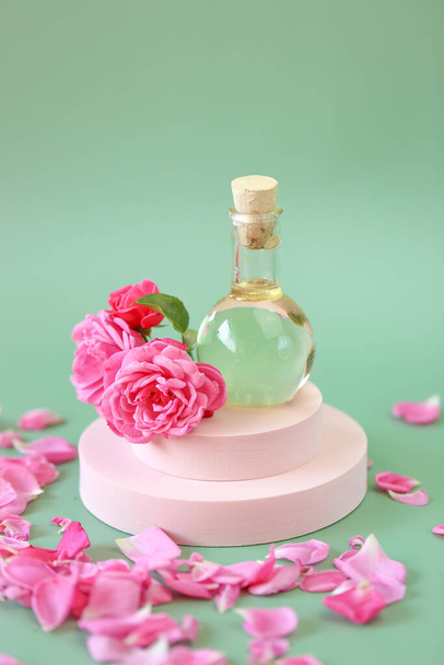Růže esenciální olej ve skleněné láhvi v růžových květinách na růžovém pódiu na zeleném pozadí.Aromaterapie a kosmetika. přírodní růžový oleje.Organická kosmetika - Fotografie, Obrázek