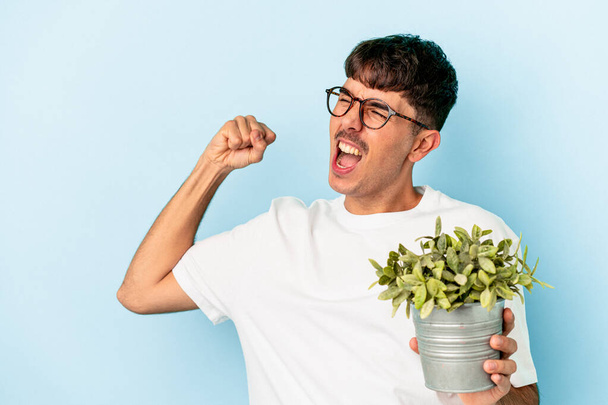 Jeune homme métis tenant une plante isolée sur fond bleu levant le poing après une victoire, concept gagnant. - Photo, image