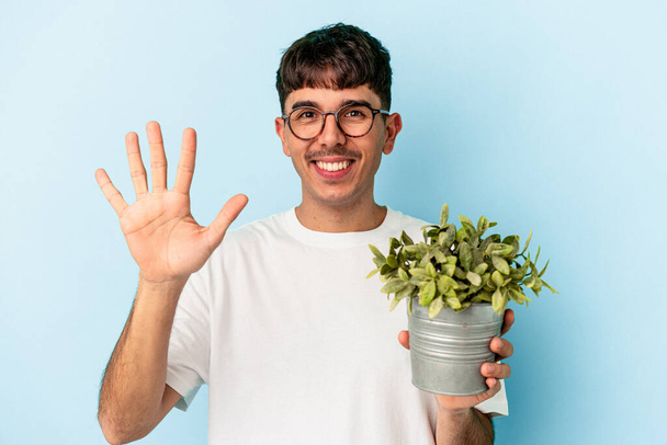 Νεαρός άνδρας μικτής φυλής κρατώντας ένα φυτό απομονωμένο σε μπλε φόντο χαμογελώντας χαρούμενα δείχνει τον αριθμό πέντε με τα δάχτυλα. - Φωτογραφία, εικόνα