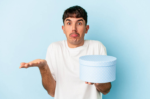 Young mixed race man holding a box present isolated on blue background shrugs shoulders and open eyes confused. - Foto, Bild