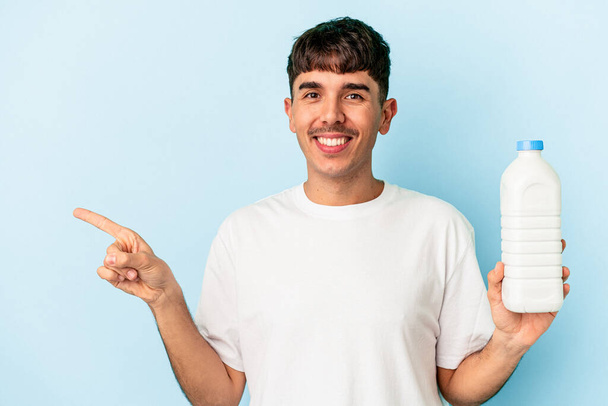 Jonge gemengde ras man met een fles melk geïsoleerd op blauwe achtergrond glimlachend en wijzend opzij, toont iets op lege ruimte. - Foto, afbeelding