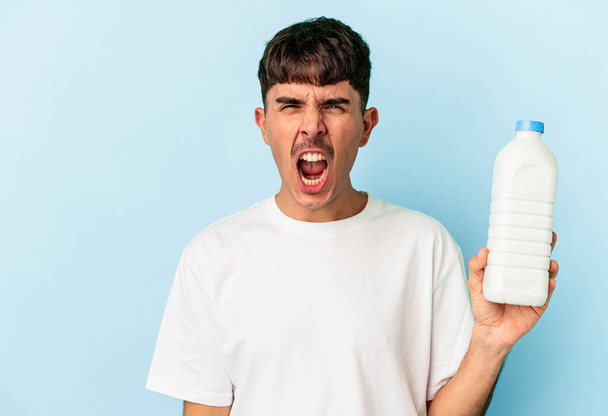 Jonge gemengde ras man met een fles melk geïsoleerd op blauwe achtergrond schreeuwen zeer boos en agressief. - Foto, afbeelding