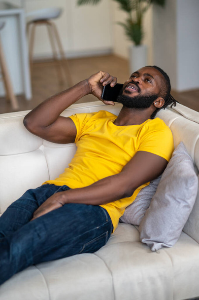 Dark-skinned man talking on smartphone lying on couch - Fotó, kép