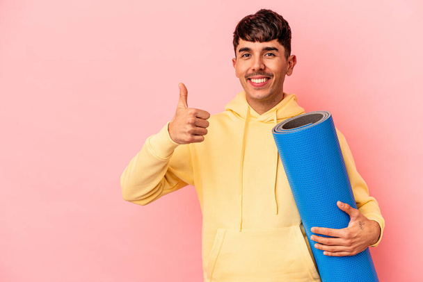 Молодой человек смешанной расы держит спортивный коврик на розовом фоне улыбаясь и поднимая большой палец вверх - Фото, изображение
