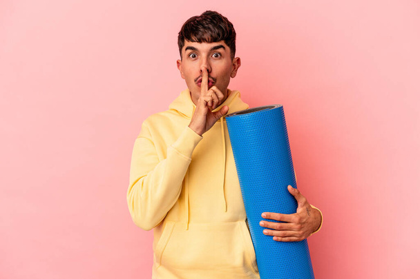 Joven hombre de raza mixta sosteniendo una alfombra deportiva aislada sobre fondo rosa manteniendo un secreto o pidiendo silencio. - Foto, Imagen