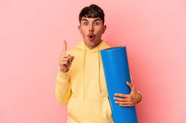 Joven hombre de raza mixta sosteniendo una alfombra deportiva aislada sobre fondo rosa que tiene una gran idea, concepto de creatividad. - Foto, Imagen