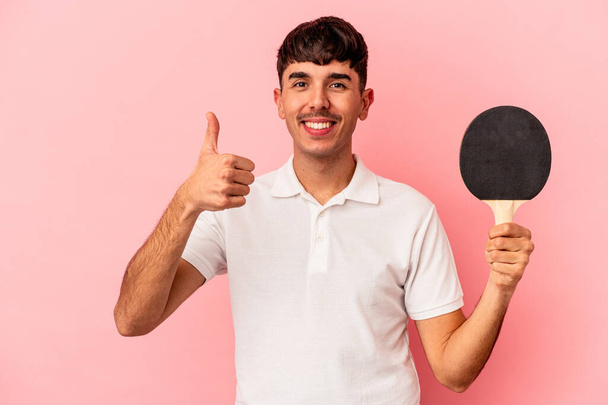 Jeune homme métis tenant une raquette de ping-pong isolé sur fond rose souriant et levant le pouce vers le haut - Photo, image