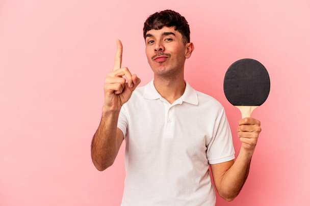 Jeune homme métis tenant une raquette de ping-pong isolé sur fond rose montrant numéro un avec doigt. - Photo, image