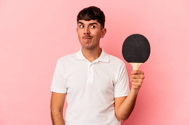 Jeune homme métis tenant une raquette de ping-pong isolé sur fond rose confus, se sent douteux et incertain. - Photo, image