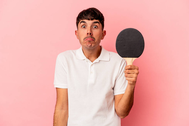 Jeune homme métis tenant une raquette de ping-pong isolé sur fond rose haussant les épaules et les yeux ouverts confus. - Photo, image