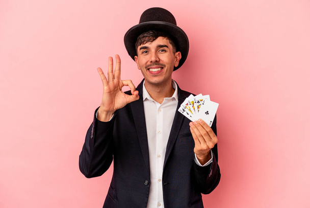 Молодой кавказский волшебник с волшебными карточками на белом фоне, веселый и уверенный в себе, показывающий нормальный жест. - Фото, изображение