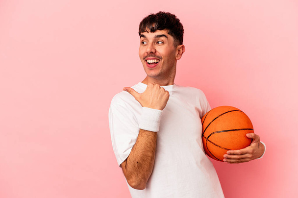 Joven hombre de raza mixta jugando baloncesto aislado en puntos de fondo rosa con el dedo pulgar lejos, riendo y sin preocupaciones. - Foto, Imagen