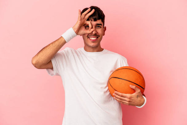 Nuori mixed race mies pelaa koripalloa eristetty vaaleanpunainen tausta innoissaan pitää ok ele silmään. - Valokuva, kuva