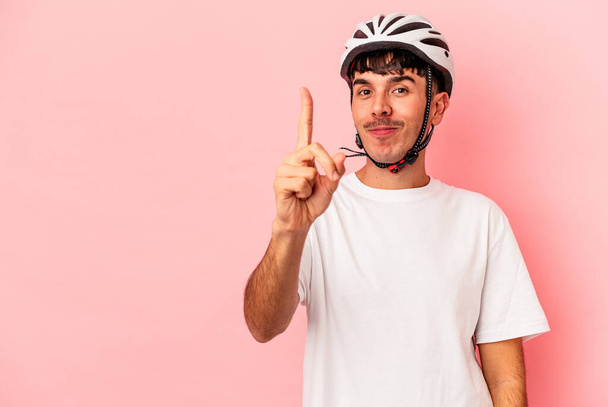 Junger Mischlingsrennfahrer mit Helm auf rosa Hintergrund zeigt Nummer eins mit Finger. - Foto, Bild