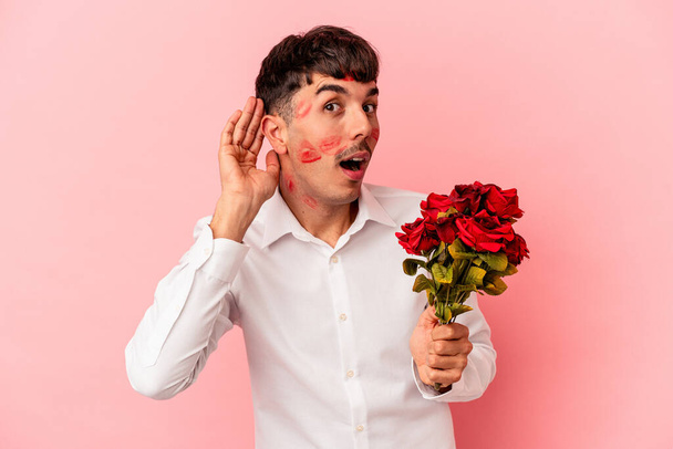Junger Mann mit gemischter Rasse hält einen Blumenstrauß auf rosa Hintergrund und versucht, einem Klatsch und Tratsch zuzuhören. - Foto, Bild