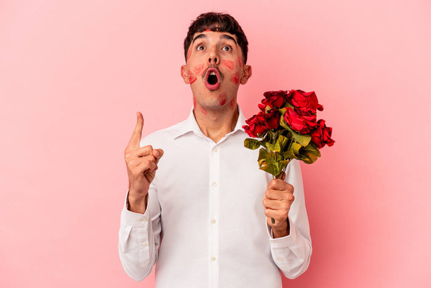 Giovane uomo razza mista in possesso di un mazzo di fiori isolati su sfondo rosa che punta verso l'alto con la bocca aperta. - Foto, immagini