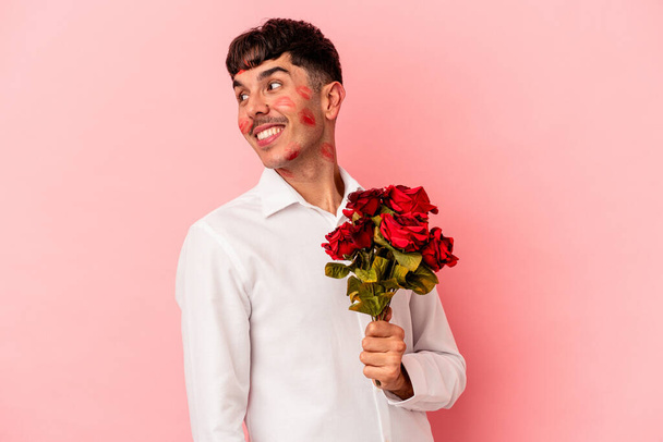 Young mixed race man holding a bouquet of flowers isolated on pink background looks aside smiling, cheerful and pleasant. - Foto, imagen
