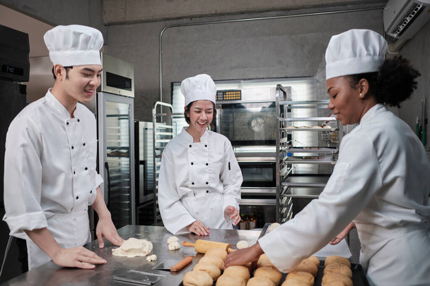 Professional gourmet team, three young chefs in white cook uniforms and aprons knead pastry dough and eggs, prepare bread, and fresh bakery food, baking in oven at stainless steel restaurant kitchen. - Foto, afbeelding