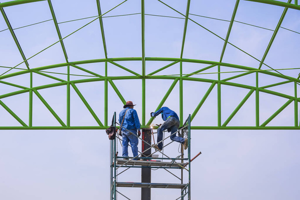 Capataz asiático y trabajador de la construcción en andamios están trabajando para construir la estructura del techo de metal de la construcción industrial en el sitio de construcción - Foto, Imagen