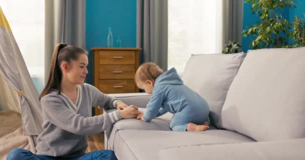 Egy gondoskodó anya ül a szőnyegen a kényelmes nappaliban és nézi, ahogy a fia ül rajta. - Felvétel, videó