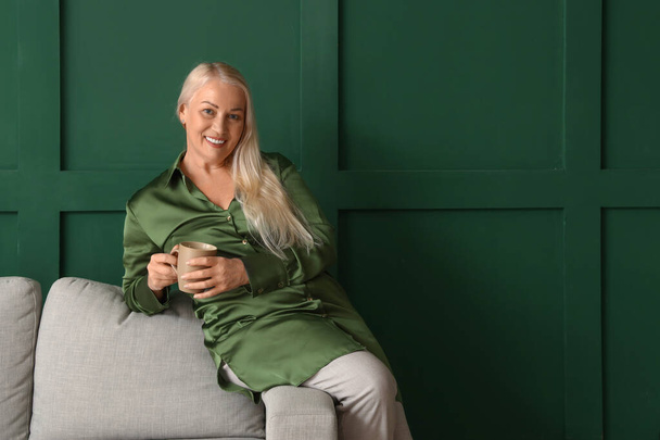 Зріла жінка п'є чай, сидячи на дивані біля зеленої стіни
 - Фото, зображення