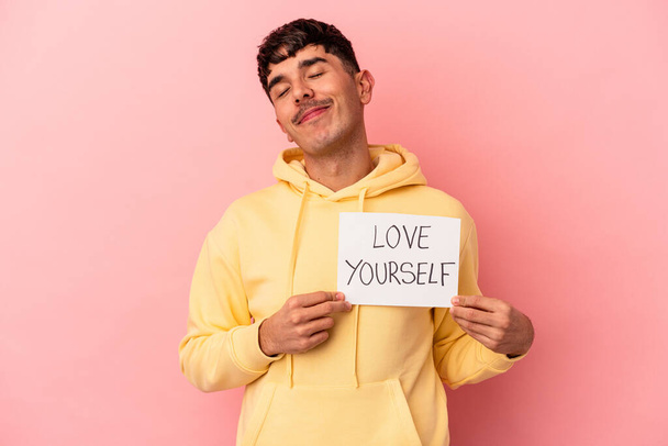 Молодой человек смешанной расы держит любовь себя плакат изолирован на розовом фоне - Фото, изображение