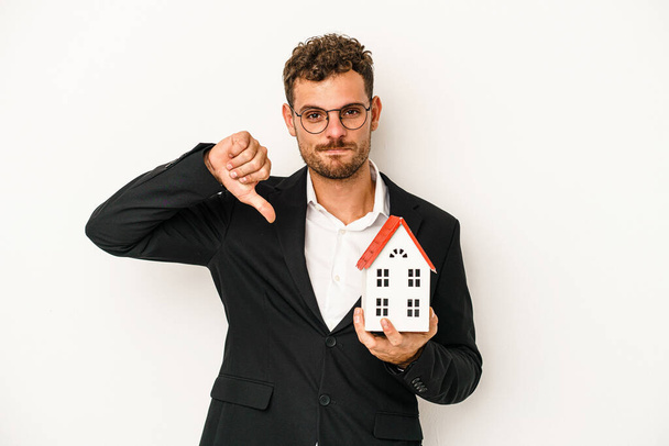 Young caucasian real estate holding a toy home isolated on white background showing a dislike gesture, thumbs down. Disagreement concept. - Fotoğraf, Görsel