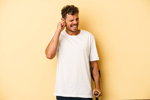 Молодий білий чоловік тримає череп ізольований на жовтому тлі, покриваючи вуха руками
. - Фото, зображення