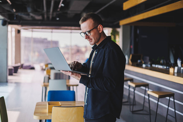 Άντρας επιχειρηματίας με μαύρο πουκάμισο και γυαλιά στέκεται στο γραφείο με φορητό υπολογιστή στο χέρι, επιτυχημένη εταιρική αφεντικό αισθάνεται καλά από πλούσιο τρόπο ζωής - Φωτογραφία, εικόνα