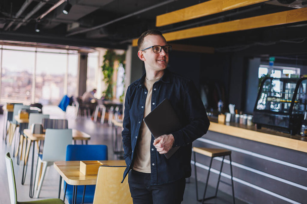 Male entrepreneur in black shirt and glasses standing in office with laptop in hand, successful corporate boss feeling good from rich lifestyle - Photo, Image