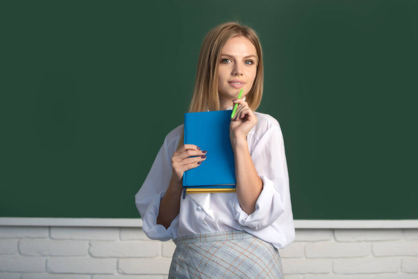 Portrait of young cute blonde female college student studying in classroom on class with blackboard background. - 写真・画像
