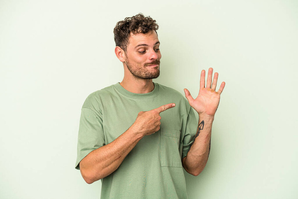 Νεαρός καυκάσιος άνδρας απομονωμένος σε πράσινο φόντο χαμογελώντας χαρούμενος δείχνοντας το νούμερο πέντε με τα δάχτυλα. - Φωτογραφία, εικόνα