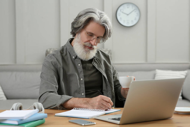Homem de meia idade com laptop, notebook e xícara de bebida aprendendo na mesa dentro de casa - Foto, Imagem