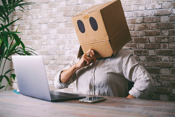 Home worker with paper box on his head. Anonymous people use laptop computer. Concept of doubts and problems with privacy online web protection. Communication and connection online - 写真・画像