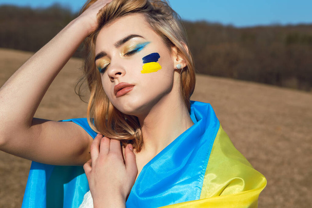 Portrait of woman wrapped in ukrainian yellow blue flag. National symbol of Ukraine on cheek. Patriotic lady with makeup walking in the field. Stand with Ukraine, support and solidarity - Фото, зображення
