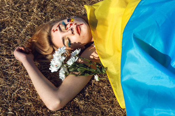 Stand with Ukraine! Woman wrapped in ukrainian yellow blue flag lying on the ground. National symbol of Ukraine. Lady with blood makeup hold chamomile flowers. Support and solidarity.  - Fotografie, Obrázek