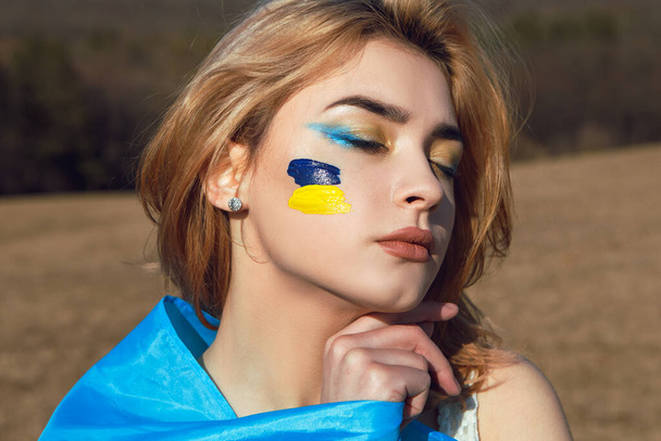 Ritratto di donna avvolto nella bandiera blu gialla ucraina. Simbolo nazionale dell'Ucraina sulla guancia. Signora patriottica truccata che cammina sul campo. Sostegno all'Ucraina, sostegno e solidarietà - Foto, immagini