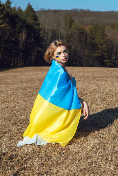 Portret van een vrouw gewikkeld in Oekraïense gele vlag. Nationaal symbool van Oekraïne op wang. Patriottische dame met make-up in het veld. Opkomen voor Oekraïne, steun en solidariteit - Foto, afbeelding